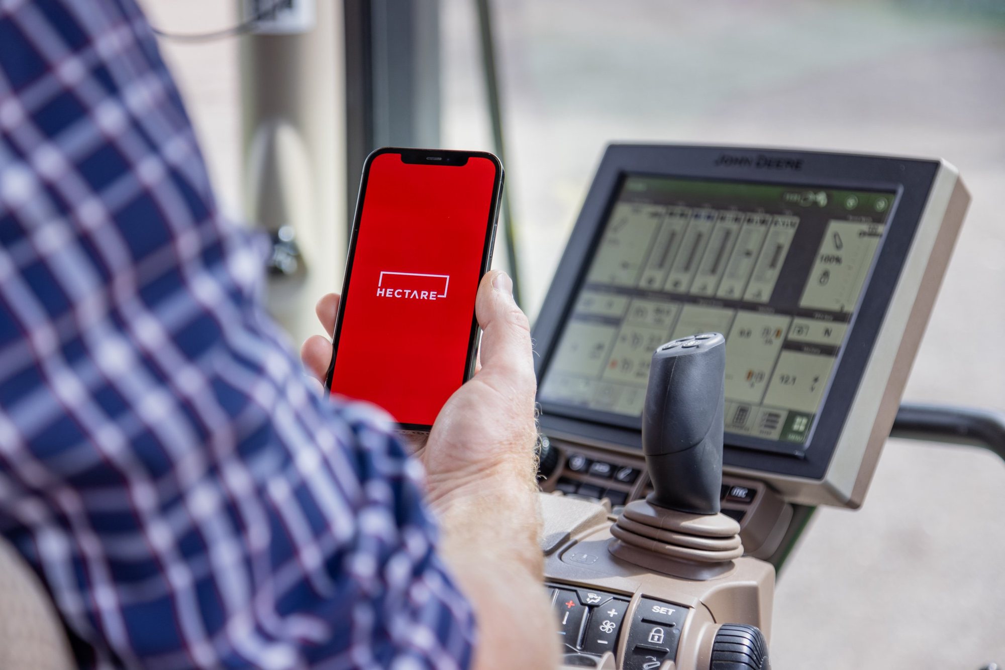 A phone showing Hectare Trading, held up beside the terminal screen in a tractor cab