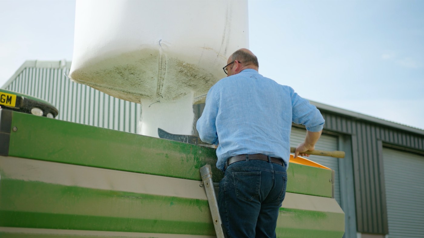 A farmer tipping grain from a bulk bag into a trailer outside the shed
